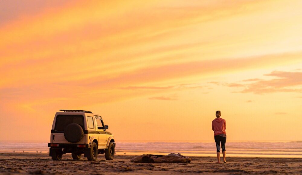 Overlanding in a Stock Vehicle Woman standing watching the sunset with her 4x4 on the beach