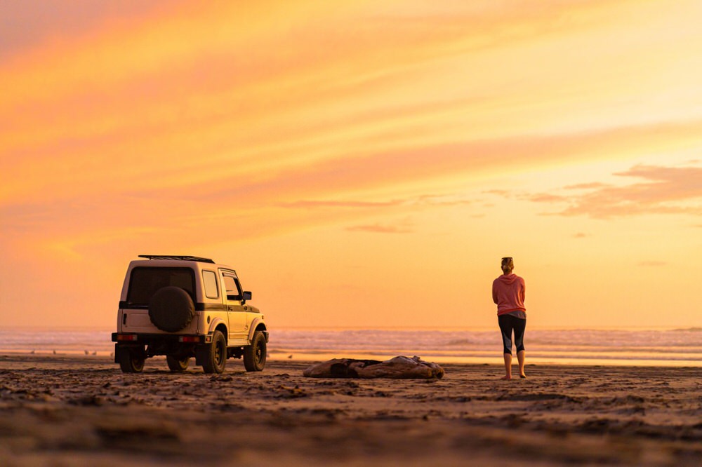 Overlanding in a Stock Vehicle Woman standing watching the sunset with her 4x4 on the beach