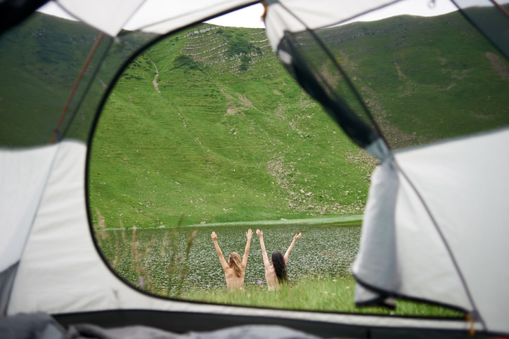 Back view of two attractive naked women lifting hands up in the air near the lake in the mountains. View from tent inside. Lifestyle concept adventure summer vacations outdoor