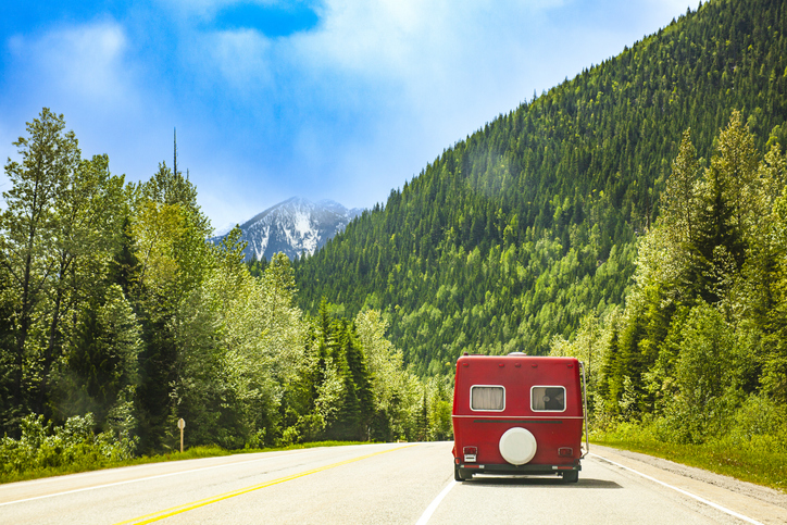 Road trip scene with vintage red caravan driving through rich green pine forest mountains and blue sky