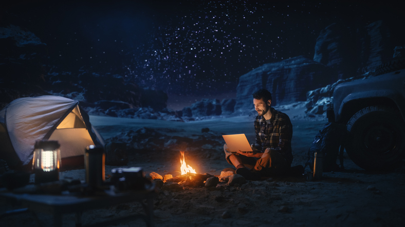 Male Traveler Sitting by Campfire Uses Laptop Computer while Tent Camping