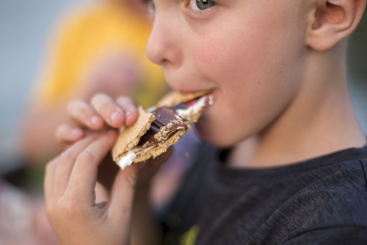 An elementary age boy takes a big bite out of a s'mores and holds it with two hands.