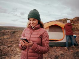 Happy young woman using smartphone during camping in mountain hill while man building tent in background