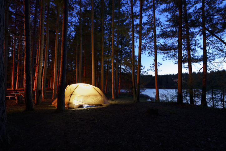 A tan tent is illuminated from within in a dark nighttime Michigan forest. There is the glow of the sky and small lake in the background. The scene also has the red light of the off camera campfire.