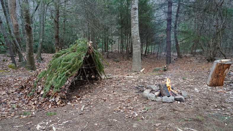 Primitive Bushcraft survival debris hut with campfire ring outside.