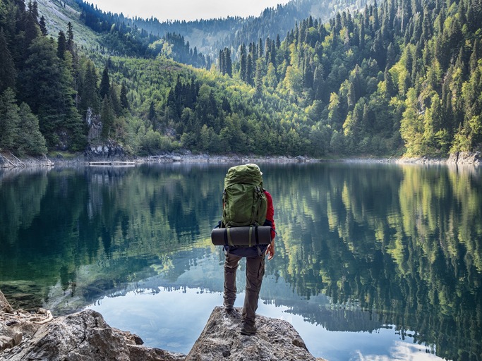Active tourist with backpack standing at mountain lake and pine wood background