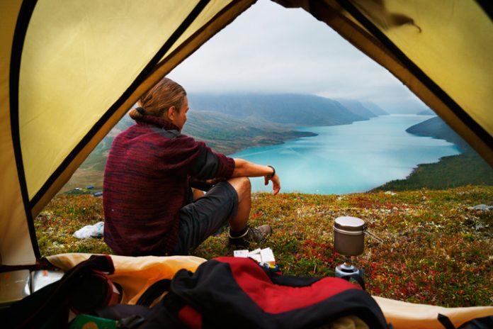 young man is sitting in front of a picturesque scenic view of lake Gjende and a remote mountain range in the Jotunheimen national park, Norway.