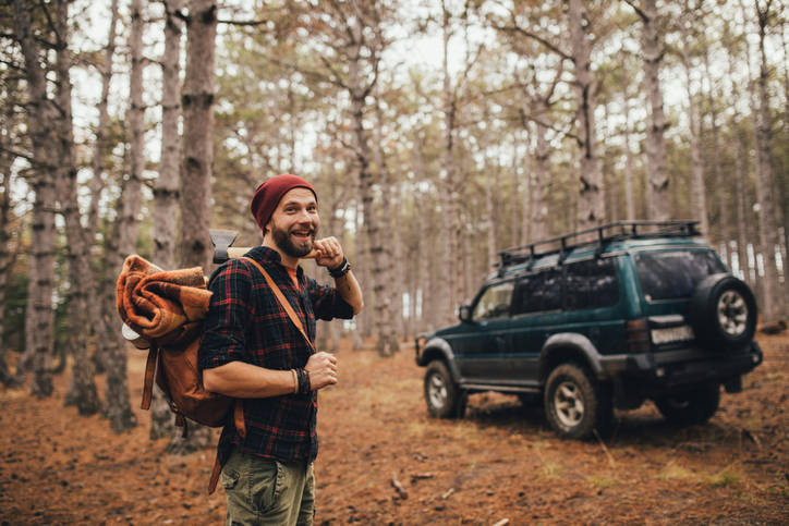 Millennial man hipster with beard wearing backpack, holding axe and firewood in a pine forest.