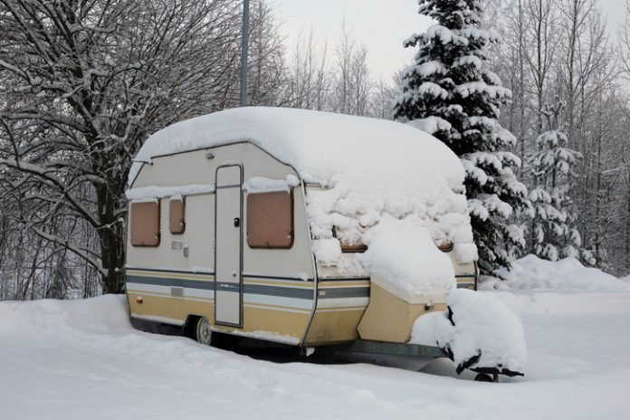 Caravan trailer in a parking lot next to a forest covered with snow during winter in Europe. The trailer is yellow and grey colored and there is snowy trees and snow around the trailer.