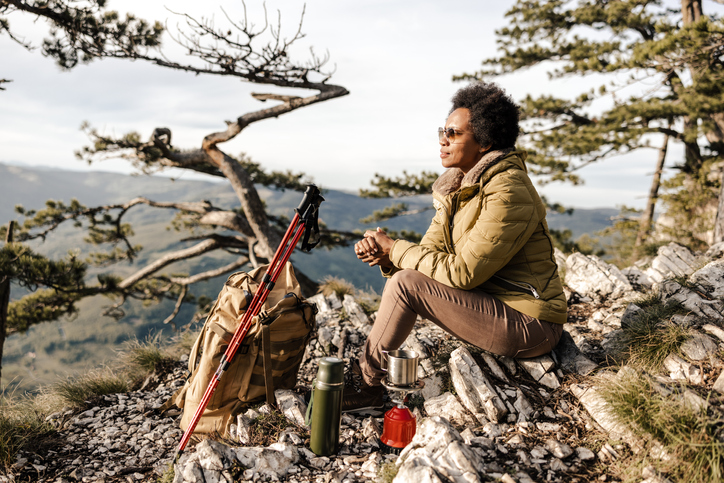 Mature black woman sitting on the top a hill and having tea break during her hike in the mountains.