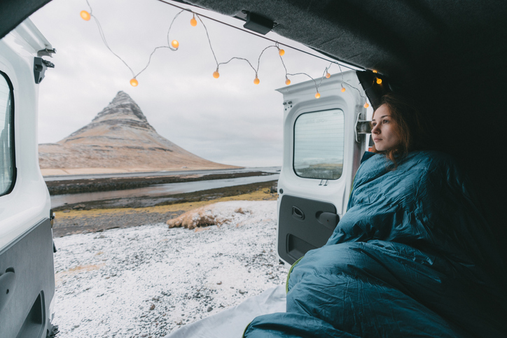 Young Caucasian woman in sleeping bag looking at Kirkjufell mountain from camper van