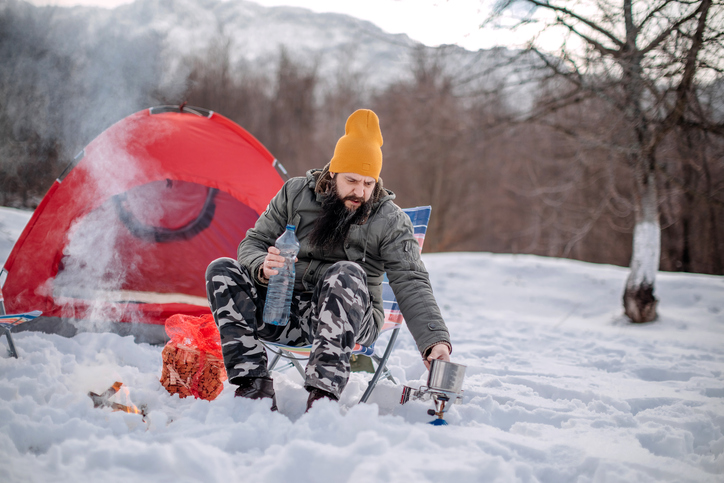 Man camping on snow with a a red tent heating water over a camp stove
