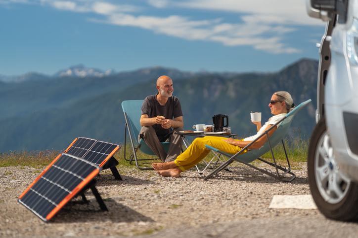Mature adult sporty couple sitting in camping chairs enjoying breakfast outdoors on their camper van vacations high up in the mountains, focus on man smiling, self supporter with solar panels