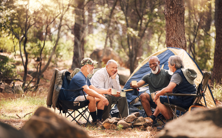 friends and camping in nature laughing for funny joke, meme or conversation by tent in forest. Group of elderly men relaxing on camp chairs with drink enjoying sunny day together in the woods
