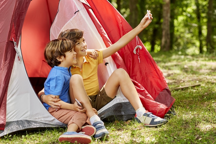 Happy brothers are taking selfie by tent. Smiling boys are camping in forest. They are spending leisure time.