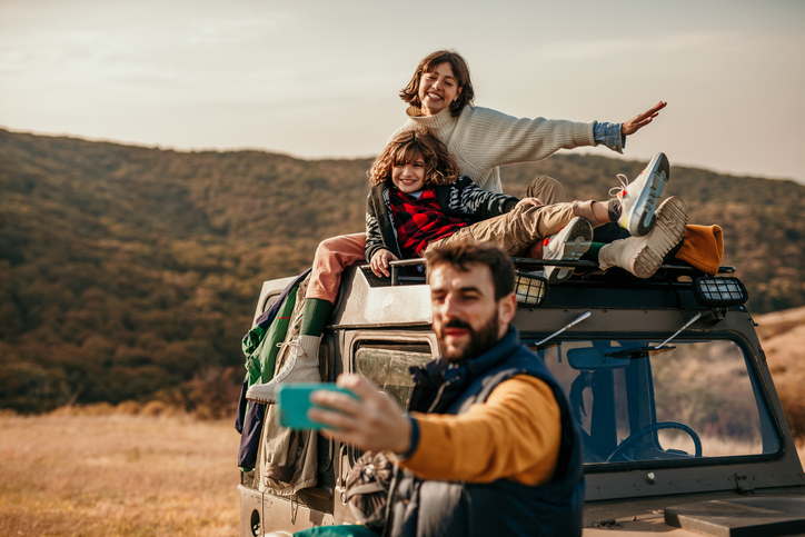 Spontaneous and beautiful photo of a cute family enjoying their adventure, camping and hiking outdoors. They’ve stopped on a beautiful landscape where a beautiful brunette, a mother, is sitting with their sun on the top of the truck, and smiling for the camera, while a father is taking a selfie of all of them. Everyone smiling, stylishly, dressed in colors