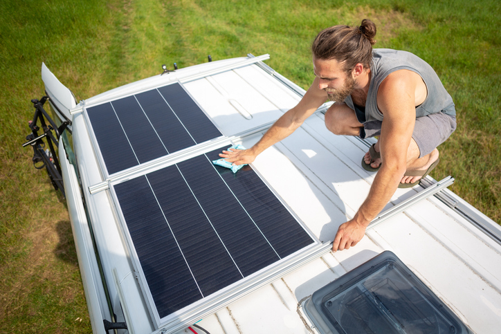 Young man on top of the roof of a camper van with solar panels. He is using a cloth to wipe clean the panels. The sun is shining and a green meadow is visible in the background.