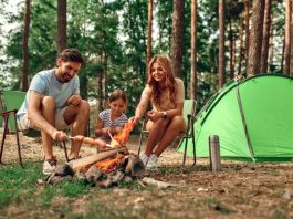 Happy family with a child on a picnic sit by the fire near the tent and grill a barbecue in a pine forest.