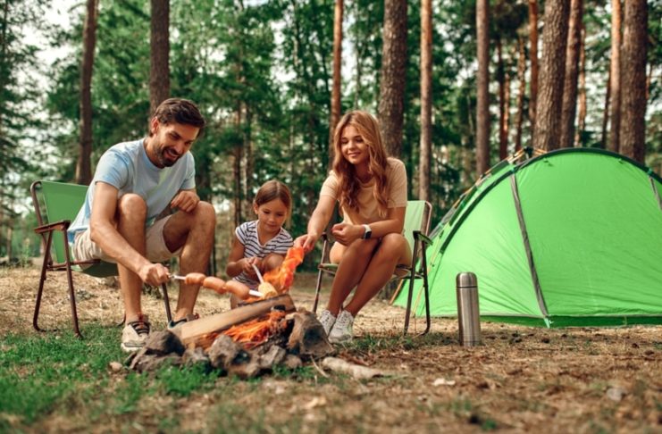 Happy family with a child on a picnic sit by the fire near the tent and grill a barbecue in a pine forest.