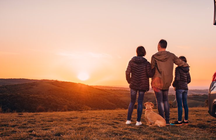 Family with small yellow dog embracing at hill and looking at sunset